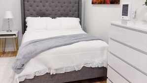 A modern bedroom with a grey tufted headboard, white bedding, a grey throw blanket, a white bedside table with a lamp, a white dresser, and a painting of a sunset on the wall. - Trackside Guesthouse w/ Pool (Clifton Park)