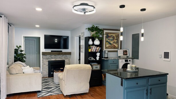 A modern living room with a fireplace, a television, a sofa, a coffee table, and a kitchen area with blue cabinets and a marble countertop. - Trackside Guesthouse w/ Pool (Clifton Park)