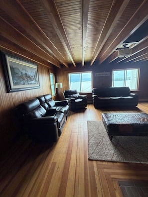 Interior de uma cabana com paredes de madeira, uma janela grande, um sofá, uma cadeira e um tapete.