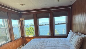 A bedroom with a large bed, wooden floors, and a view of a body of water through multiple windows.