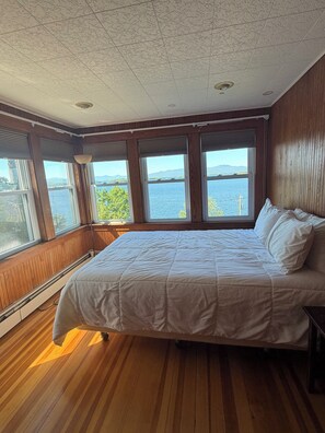 A bedroom with a large bed, wooden floors, and a view of a body of water through multiple windows. - Enjoy Breath-taking Lake and Mountains views at the Belknap Point inn House (Gilford)