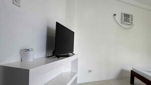 Une chambre moderne et minimaliste, dotée d’un téléviseur à écran plat posé sur un meuble blanc, d’une petite poubelle blanche et d’un lit avec un cadre en bois.