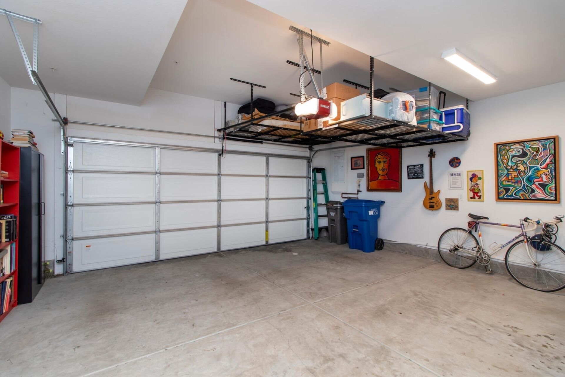 Un garage avec une porte de garage blanche, un vélo, une échelle et divers objets rangés sur des étagères.