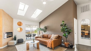 A spacious living room with a leather sofa, a coffee table, and a patterned rug. There is a fireplace, a large window, and a view of the outdoors. - Sunny Vista – Location/Views/Sleeps 8 #SVJT (Joshua Tree)