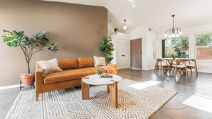 A spacious living area with a brown sofa, a coffee table, and a patterned rug. There are potted plants and a dining area with wooden chairs and a table. - Sunny Vista – Location/Views/Sleeps 8 #SVJT (Joshua Tree)