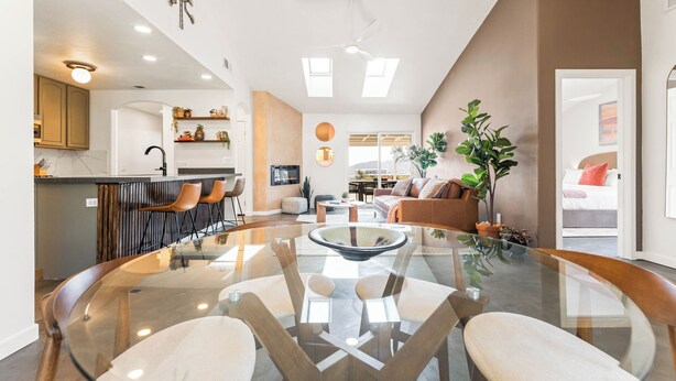 A modern living room with a glass dining table, comfortable seating, a kitchen area with bar stools, and a cozy seating area with a sofa and plants. - Sunny Vista – Location/Views/Sleeps 8 #SVJT (Joshua Tree)
