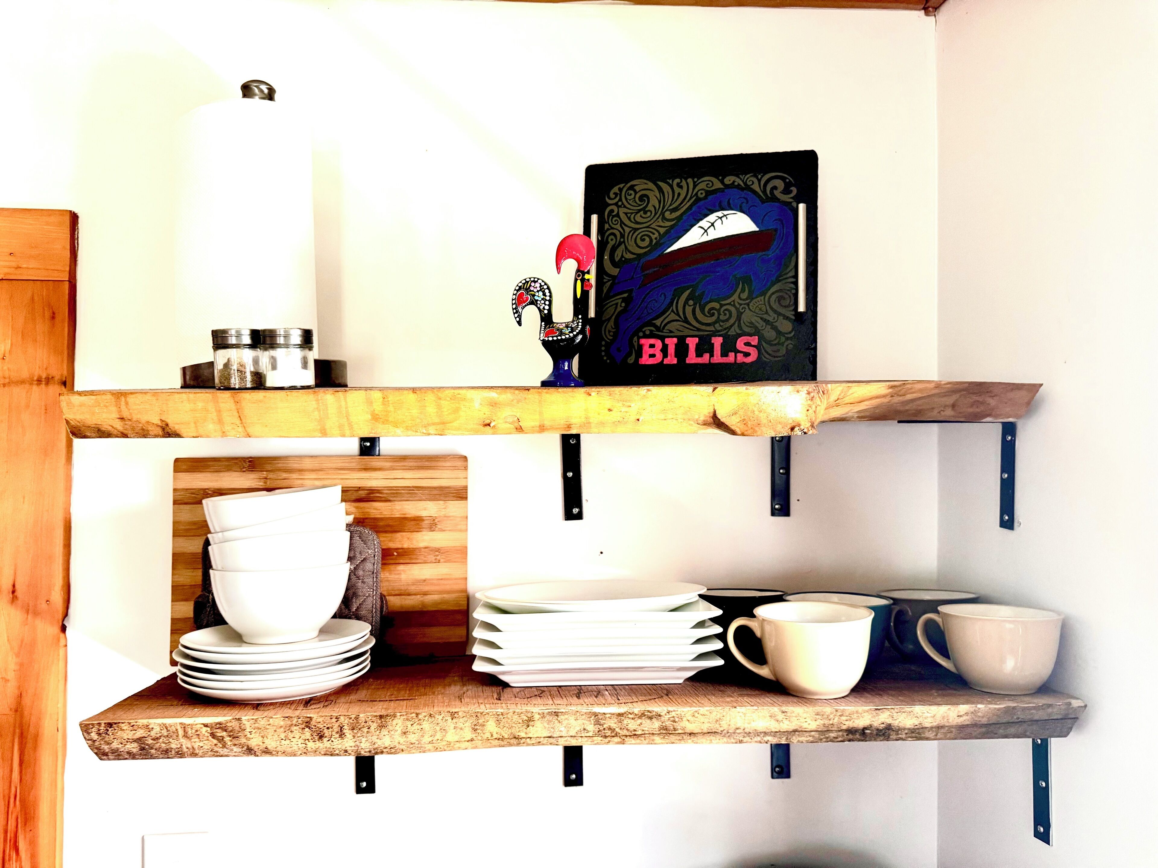 A kitchen shelf with stacked plates, a coffee cup, and a decorative item labeled "BILLS".