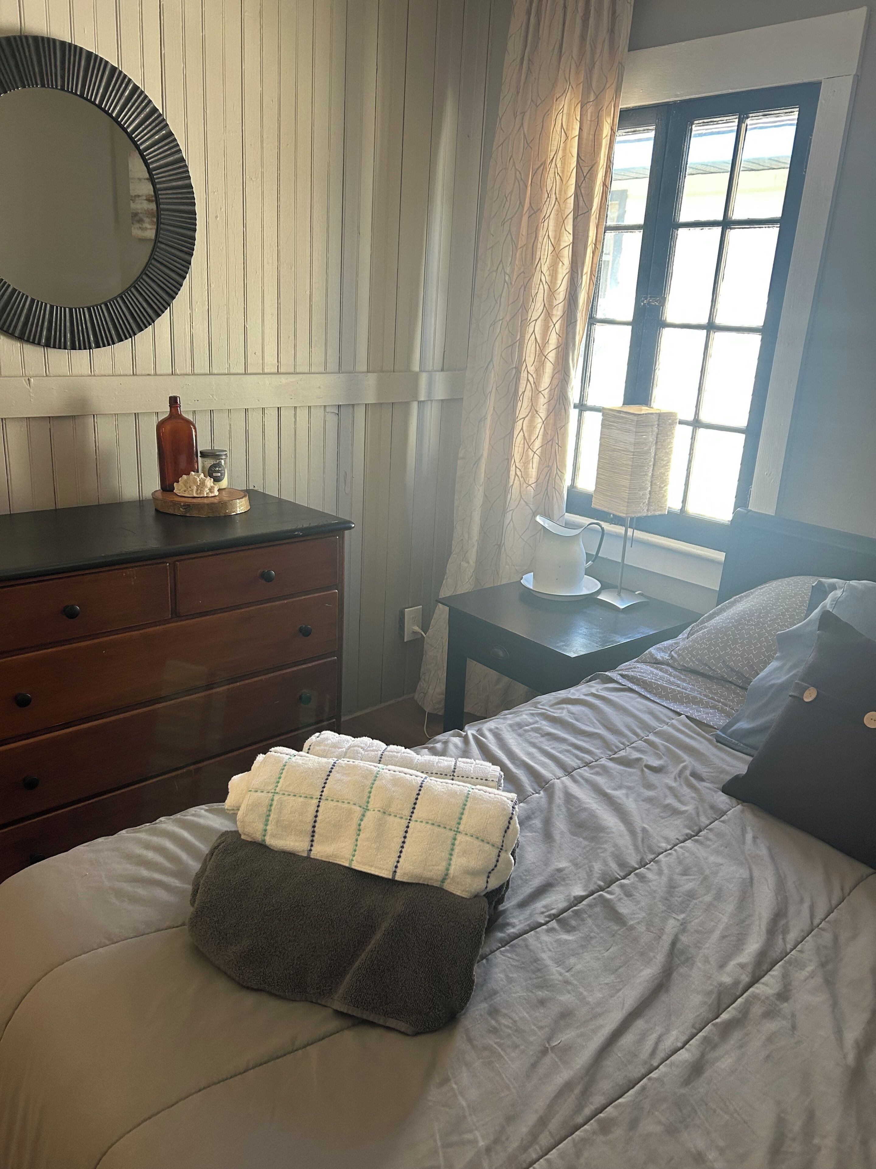 Une chambre à coucher comprenant un lit, une commode, un miroir, une fenêtre avec des rideaux et une table de chevet.