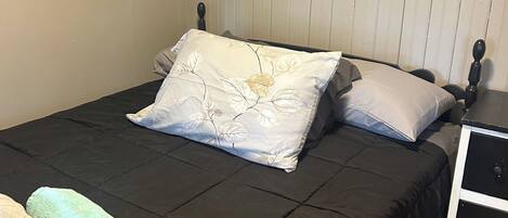 Una cama bien hecha con un cojín estampado con motivos florales, una toalla enrollada y una mesita de noche con cajón.