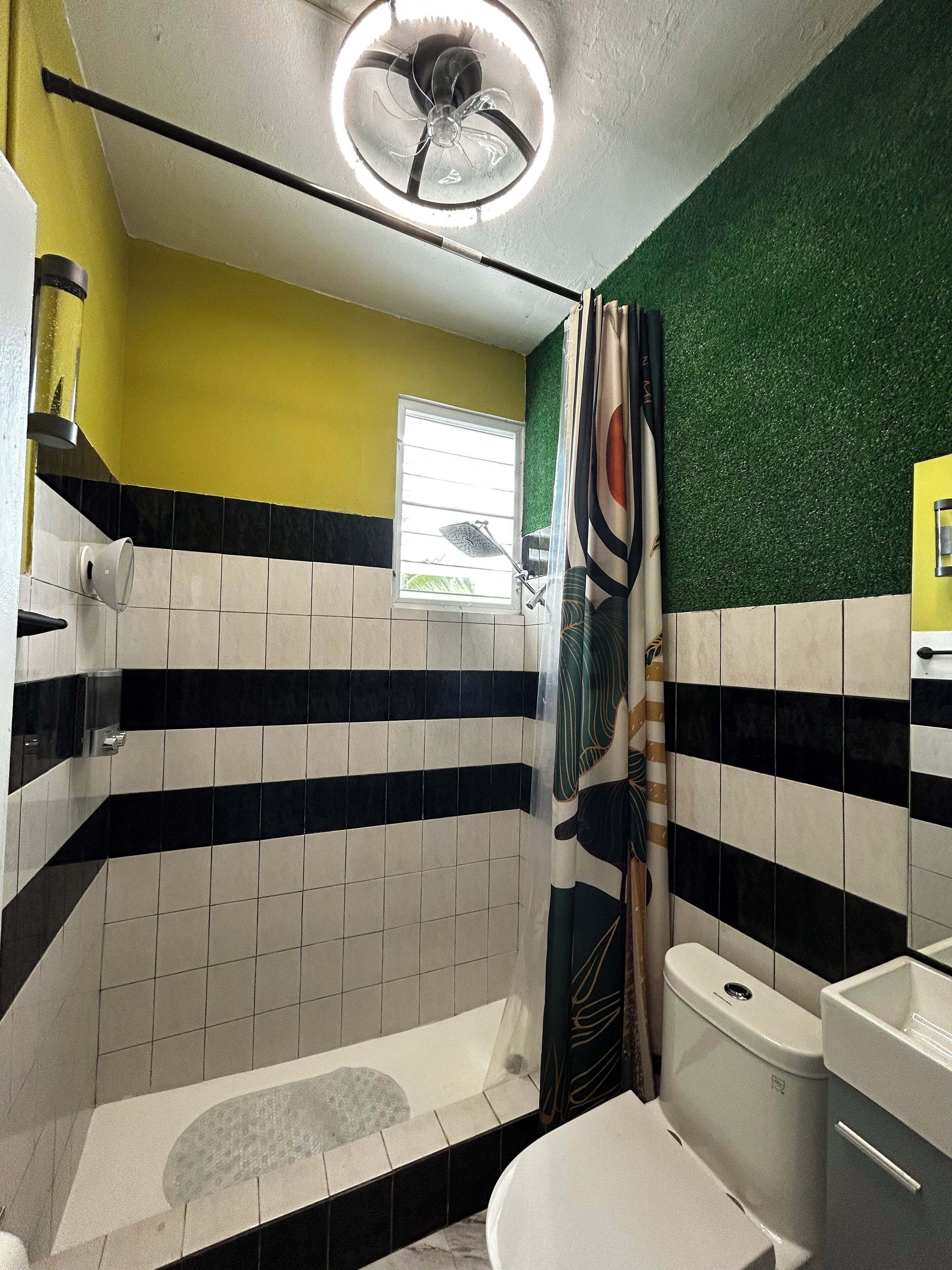 A bathroom with a shower, toilet, and sink. There is a window with blinds and a ceiling fan.