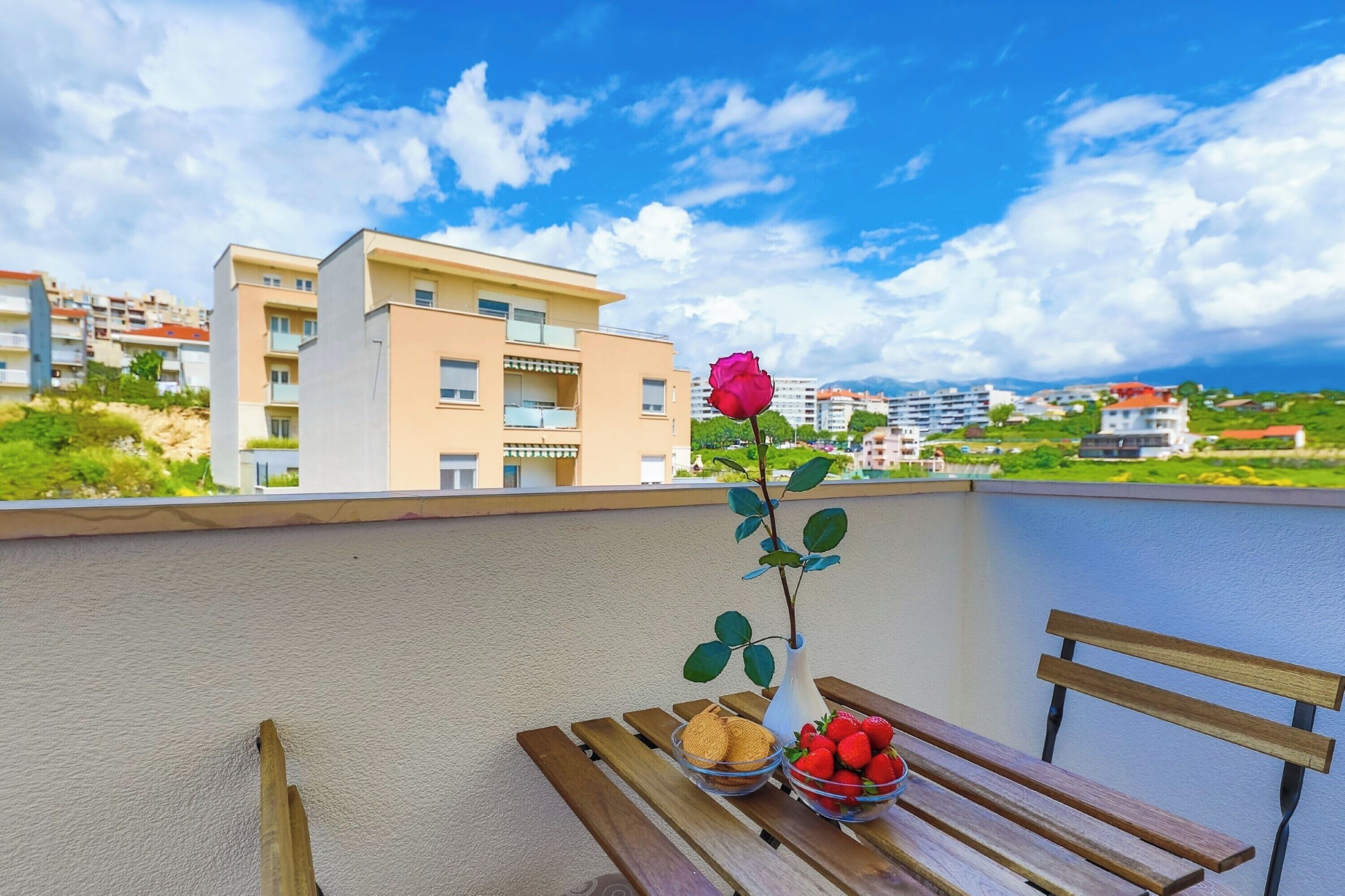 En balkon med en træbænk, en vase med en enkelt rød rose, samt skåle med jordbær og småkager.