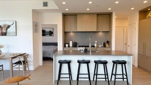 A modern kitchen with a central island, bar stools, wooden cabinets, and a dining area with a table and chairs. - New Luxury Beachfront Escape with Pool and Hottub (Kelowna)