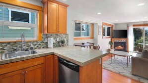 Una cocina moderna con armarios de madera, lavavajillas y salpicadero de piedra. Hay un comedor con mesa y sillas, y una chimenea en la sala de estar.