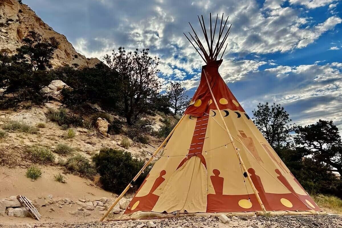 A traditional tipi with a conical structure and a circular entrance, set against a rocky landscape with sparse vegetation.
