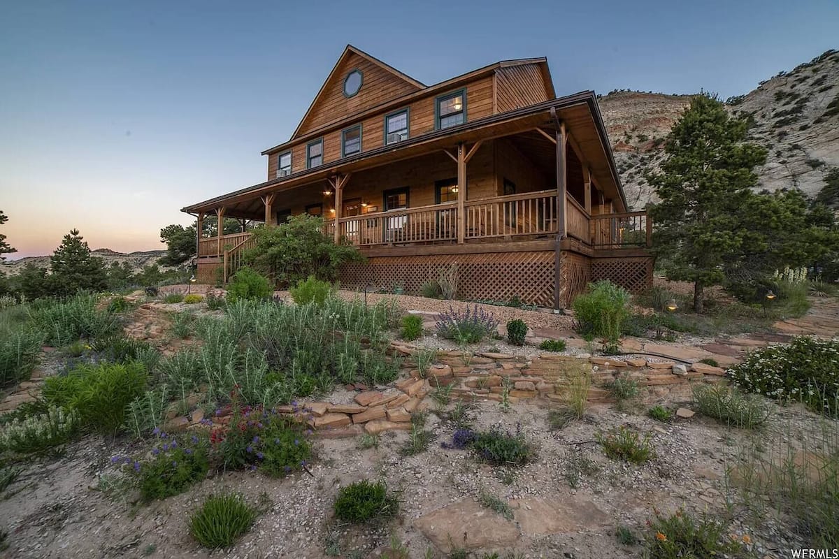 A large wooden house with a porch, surrounded by a landscaped garden with various plants and a stone pathway.