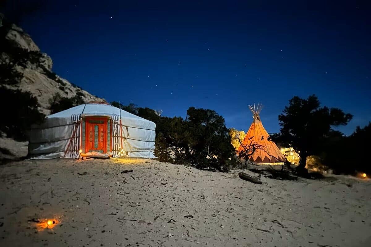 A yurt with a red door and a conical structure with lights, set against a night sky.