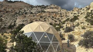 Una tienda en forma de cúpula geodésica en un paisaje desértico con vegetación escasa y colinas rocosas al fondo.
