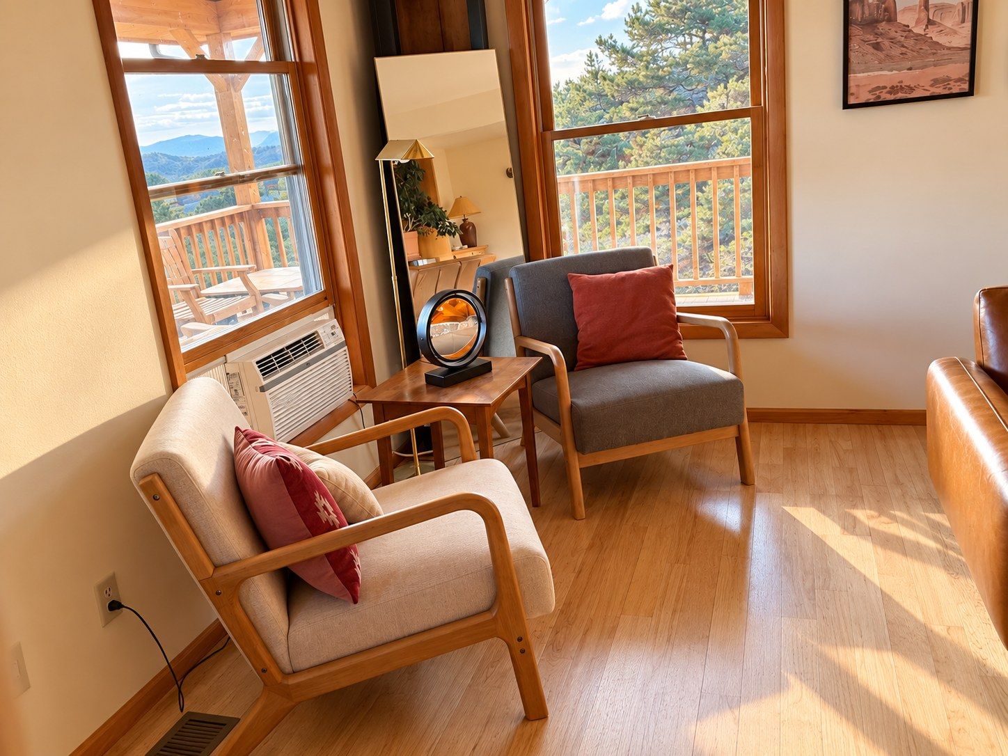 A cozy room with a wooden floor, a sofa, a chair, a table with a lamp, and a large window offering a view of trees and a balcony.