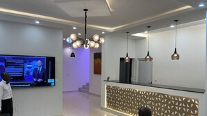 A modern hotel lobby with a large screen displaying a news broadcast, a reception desk, and a seating area with a patterned wall and pendant lighting. - Fanaseti (Conakry)