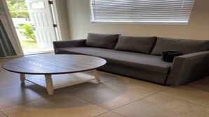 A living room with a beige sofa, a round coffee table, and a sliding glass door leading to a garden. - Charming Studio apartment in delightful Pompano Beach 1 block from the ocean (Pompano Beach)