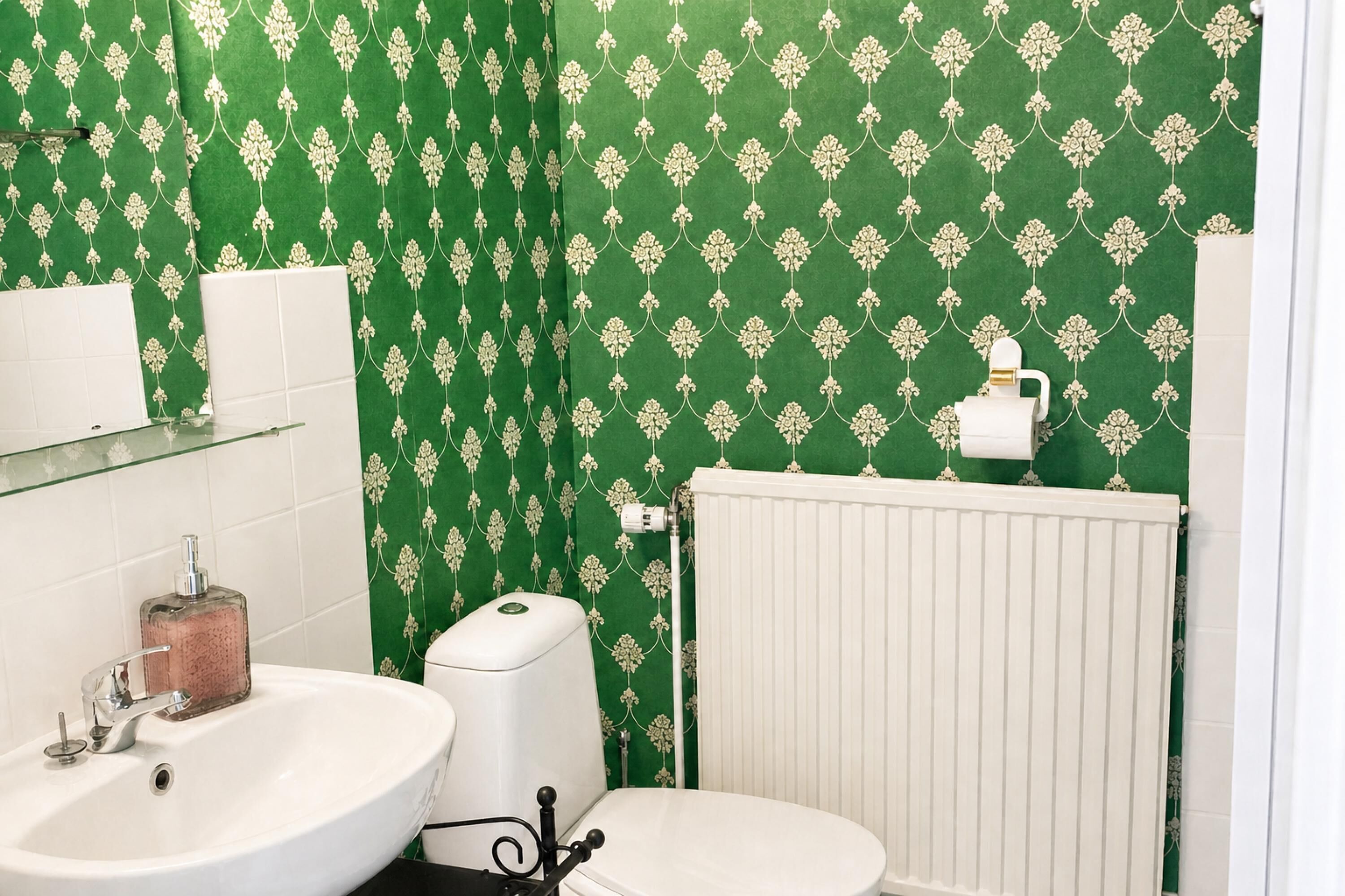 Un baño con papel tapiz verde estampado, un lavabo blanco, un inodoro y un radiador.