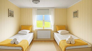 Une chambre avec deux lits simples, chacun recouvert d’une couverture jaune et agrémenté d’une serviette enroulée. On y trouve une fenêtre avec des rideaux blancs, un radiateur en dessous et un luminaire au plafond.