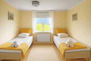 Een kamer met twee eenpersoonsbedden, elk met een gele deken en een opgerolde handdoek. Er is een raam met witte gordijnen, een radiator eronder en een plafondlamp.
