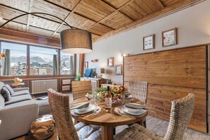 Un coin repas chaleureux avec une table en bois dressée pour deux personnes, entouré de chaises en osier, une grande fenêtre offrant une vue sur des montagnes enneigées et un plafond avec des poutres apparentes.
