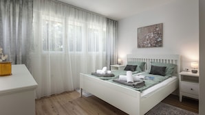 Een keurig opgemaakt slaapkamer met een bed, nachtkastjes, een bureau en een raam met dunne gordijnen.
