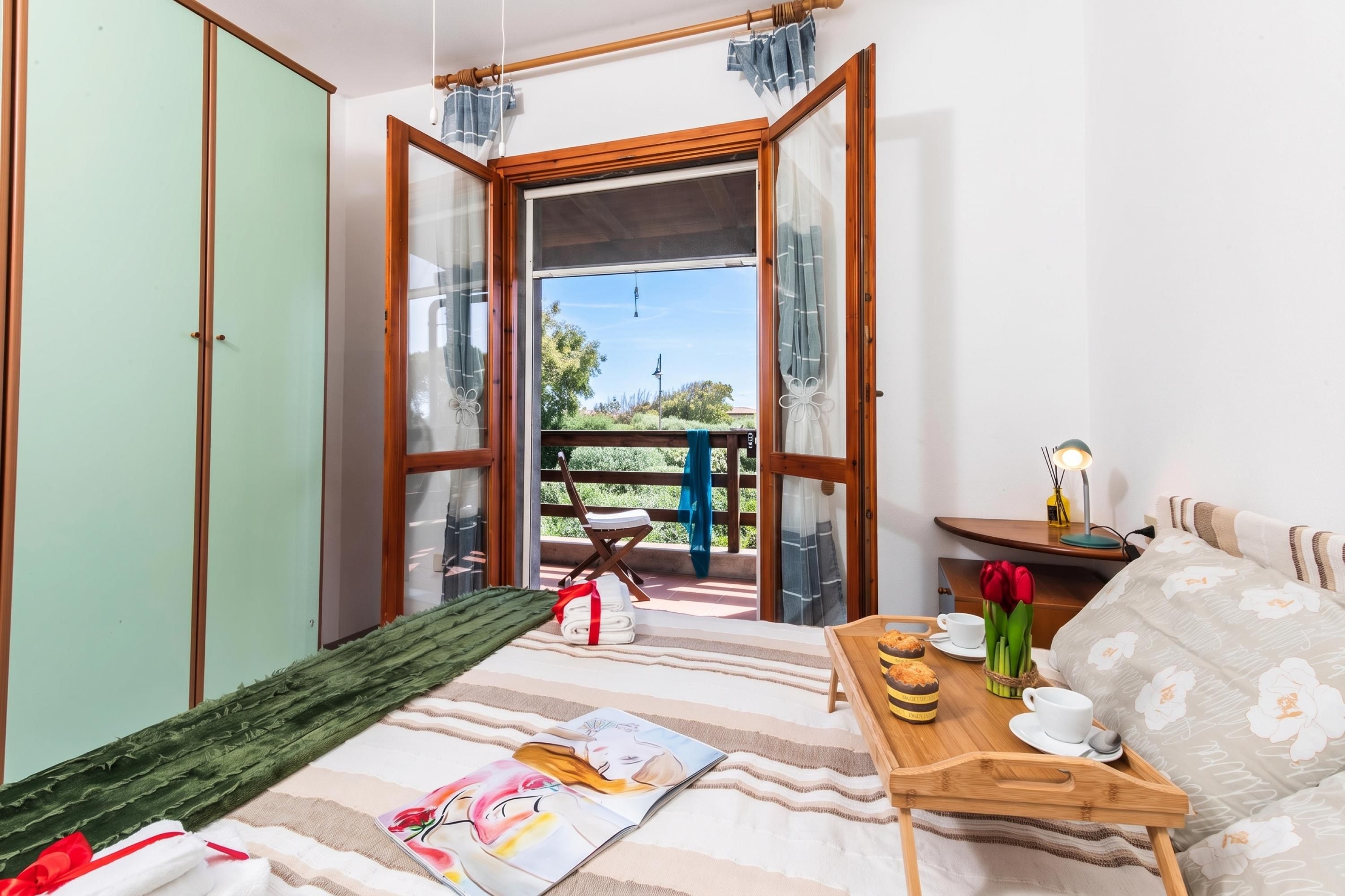 A room with a bed, a wooden side table with a cup and a book, a window with a view of greenery, and a door leading outside.