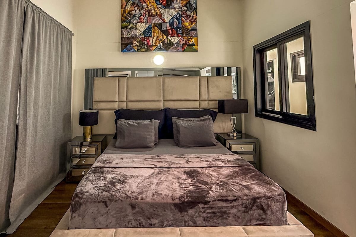 Un dormitorio con una cama grande, cabecera, dos mesitas de noche, un ventanal con vistas y un cuadro en la pared.