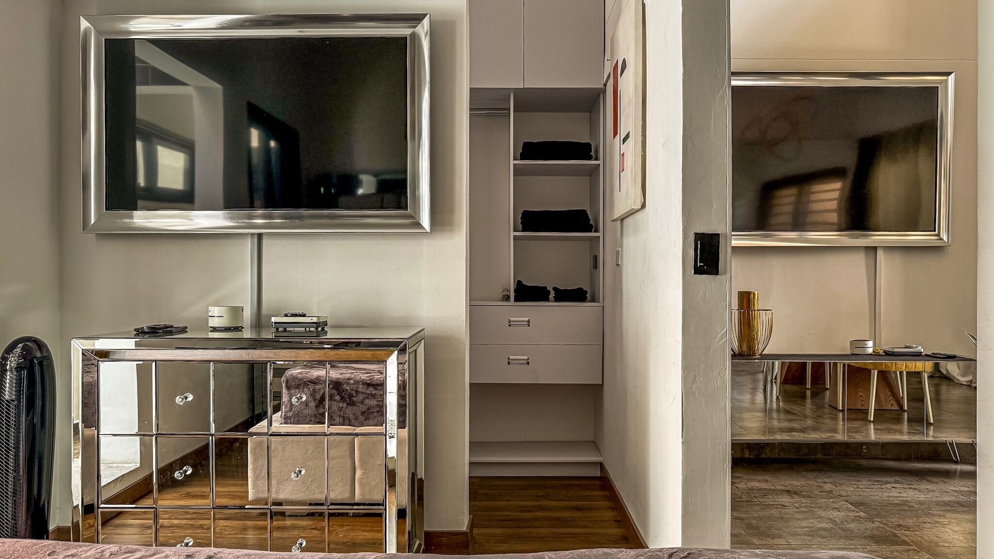 Un dormitorio moderno con un espejo grande, una mesita de noche con lámpara, un armario y una pequeña zona de estar con una mesa y sillas.