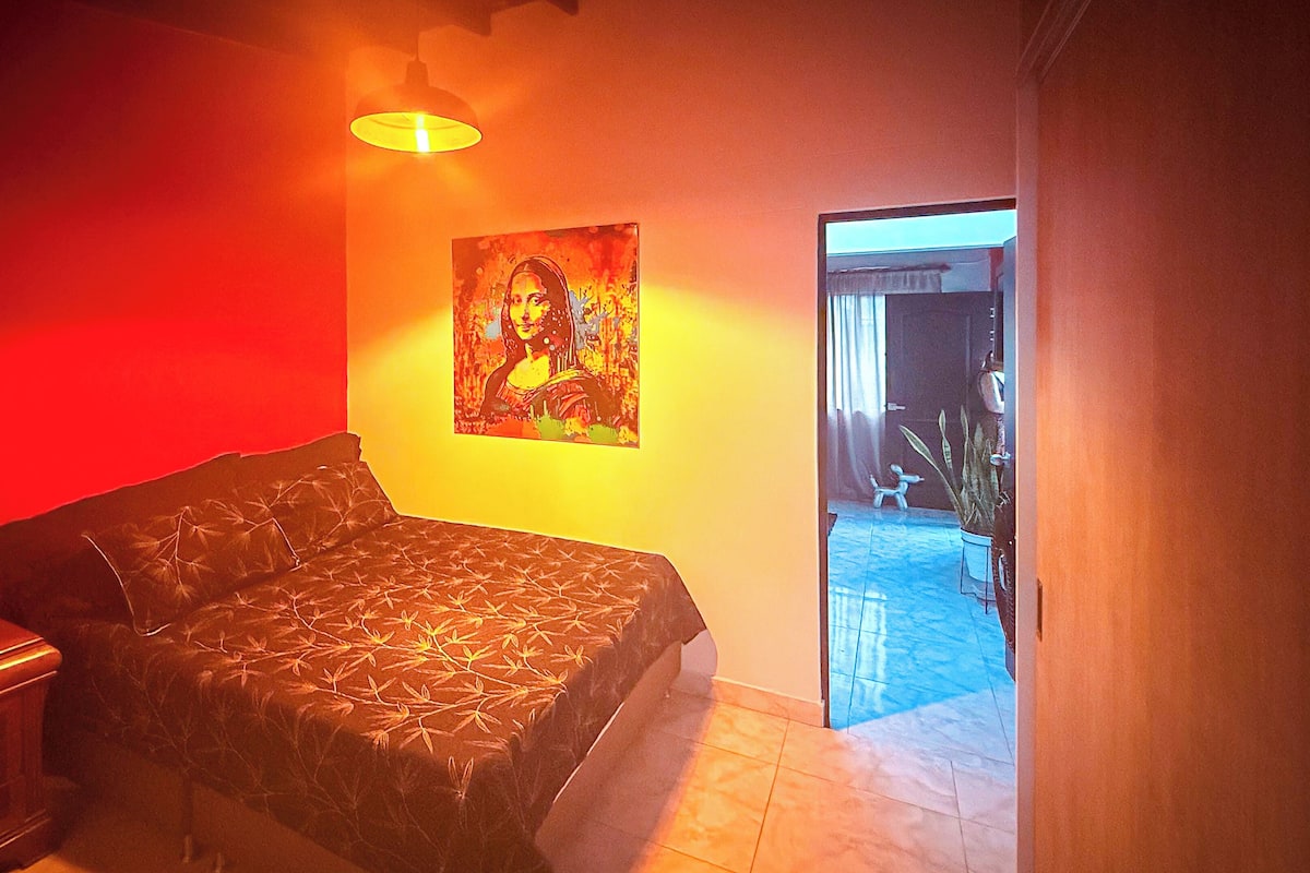 Un dormitorio con una cama, una mesita de noche, una lámpara, un cuadro en la pared y vistas al exterior.