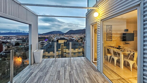 En balkong med trägolv, en glasdörr som leder till en matplats med bord och stolar, och en utsikt över en stadsbild med berg i bakgrunden.