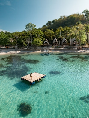 A clear turquoise sea with a wooden platform and a person standing on it. In the background, there are thatched-roof huts and lush green trees.