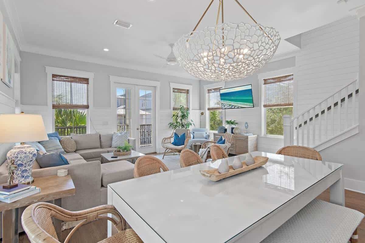 A spacious living room with a large white dining table, comfortable seating, a decorative chandelier, and a television mounted on the wall.