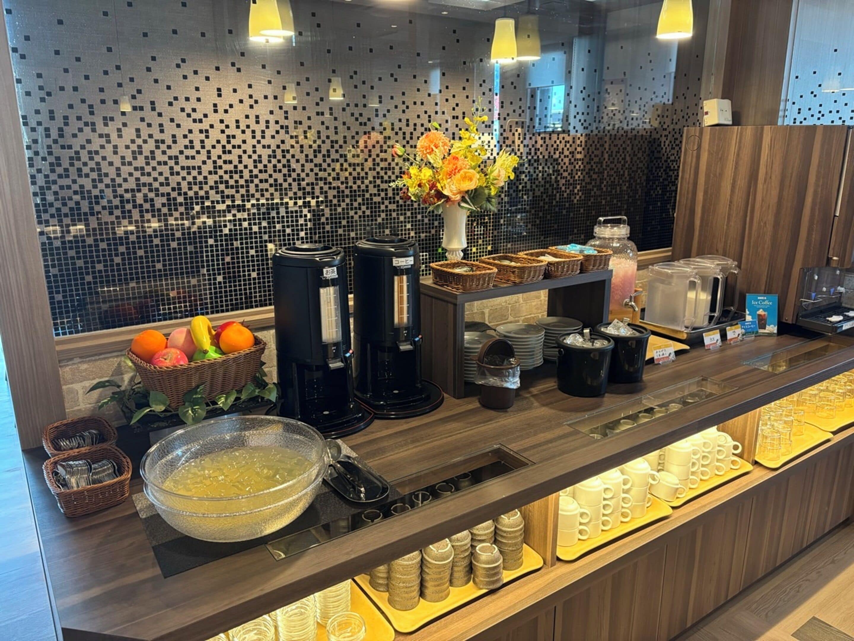 A breakfast bar with coffee machines, a fruit basket, a bowl of cereal, and various mugs and cups.