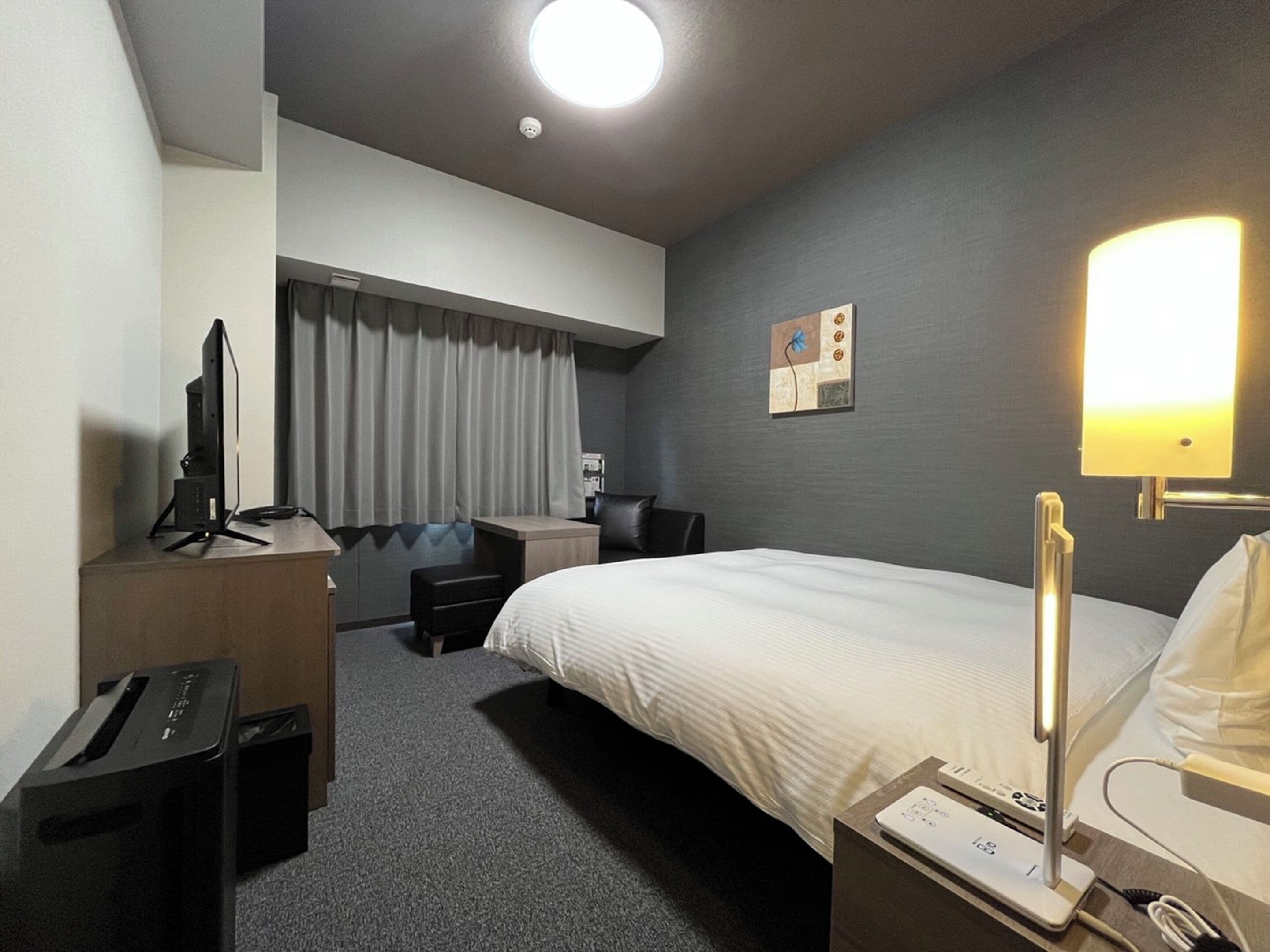 A hotel room with a bed, desk, chair, and a desk lamp.
