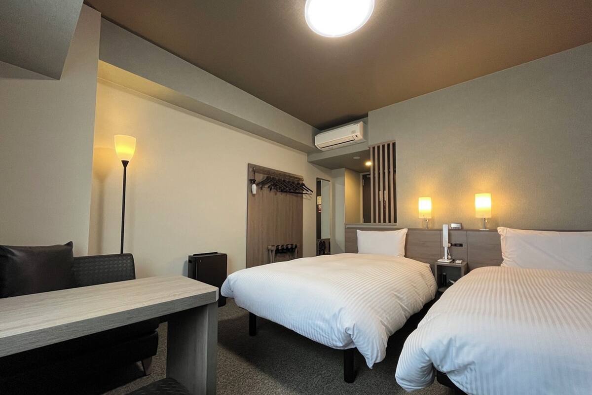 A hotel room with two beds, a desk, a chair, a lamp, and an air conditioning unit.