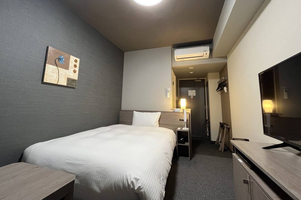 A hotel room with a bed, bedside table, lamp, TV, and an air conditioning unit.