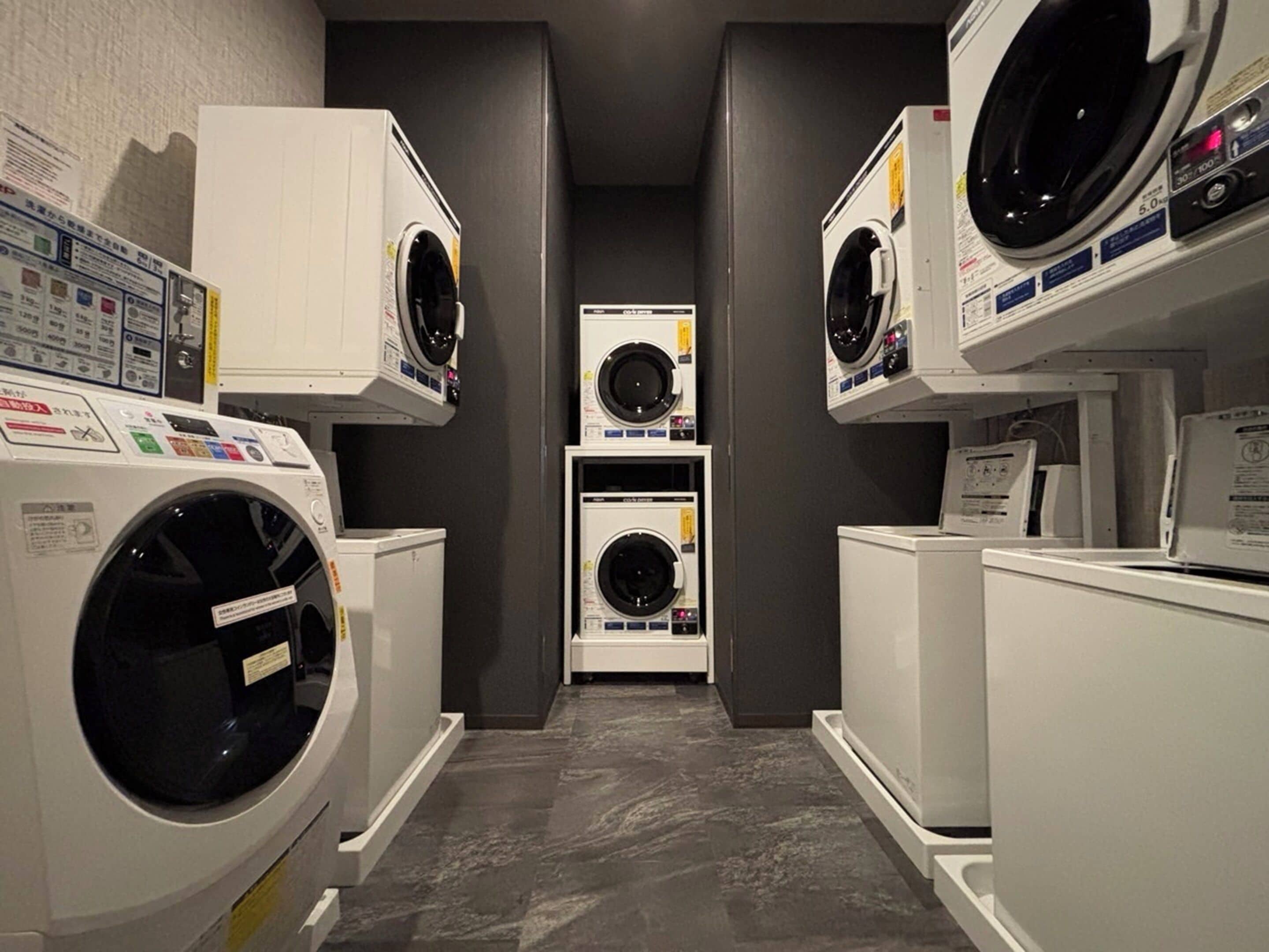 A laundry room with multiple washing machines and dryers, arranged in a narrow space with grey walls and a tiled floor.