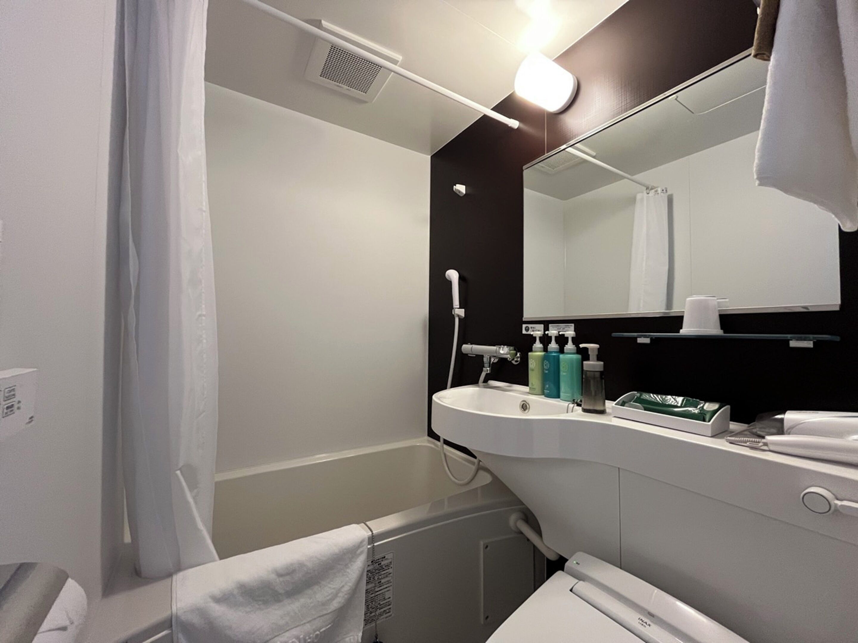 A modern bathroom with a white sink, mirror, and shower. There are toiletries on the sink and a towel hanging on the wall.