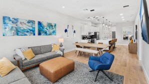 Un salon moderne avec un canapé gris, un fauteuil bleu et une table basse en bois. On y trouve également un coin repas avec une table et des chaises en bois, et une cuisine à l’arrière-plan.