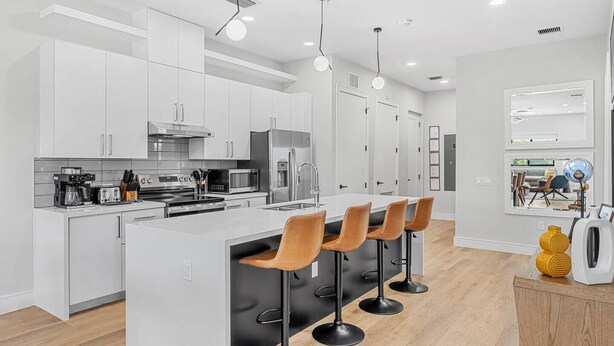 Une cuisine moderne avec des meubles blancs, un îlot central doté de tabourets de bar, des appareils en acier inoxydable et un éclairage suspendu.