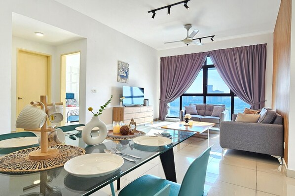 A modern living room with a glass dining table, a sofa, and a large window with purple curtains.