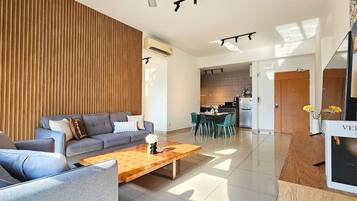 Una sala de estar moderna con un sofá gris, detalles de madera, una mesa de centro de madera y una cocina al fondo.