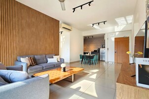 Una sala de estar moderna con un sofá gris, detalles de madera, una mesa de centro de madera y una cocina al fondo.