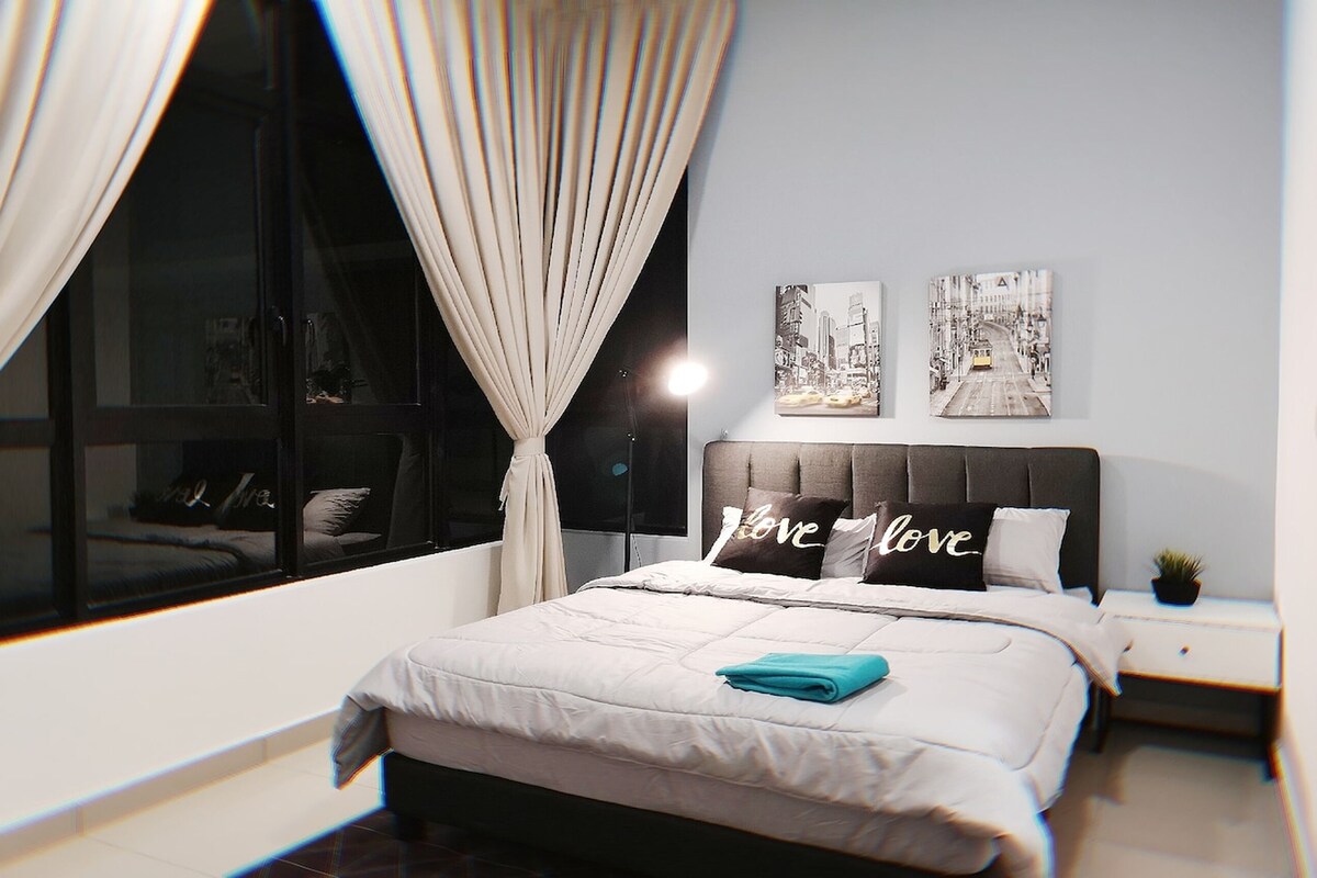 Une chambre moderne avec une grande fenêtre, un lit orné d’un coussin sur lequel est écrit le mot « Amour », et deux tableaux encadrés accrochés au mur.