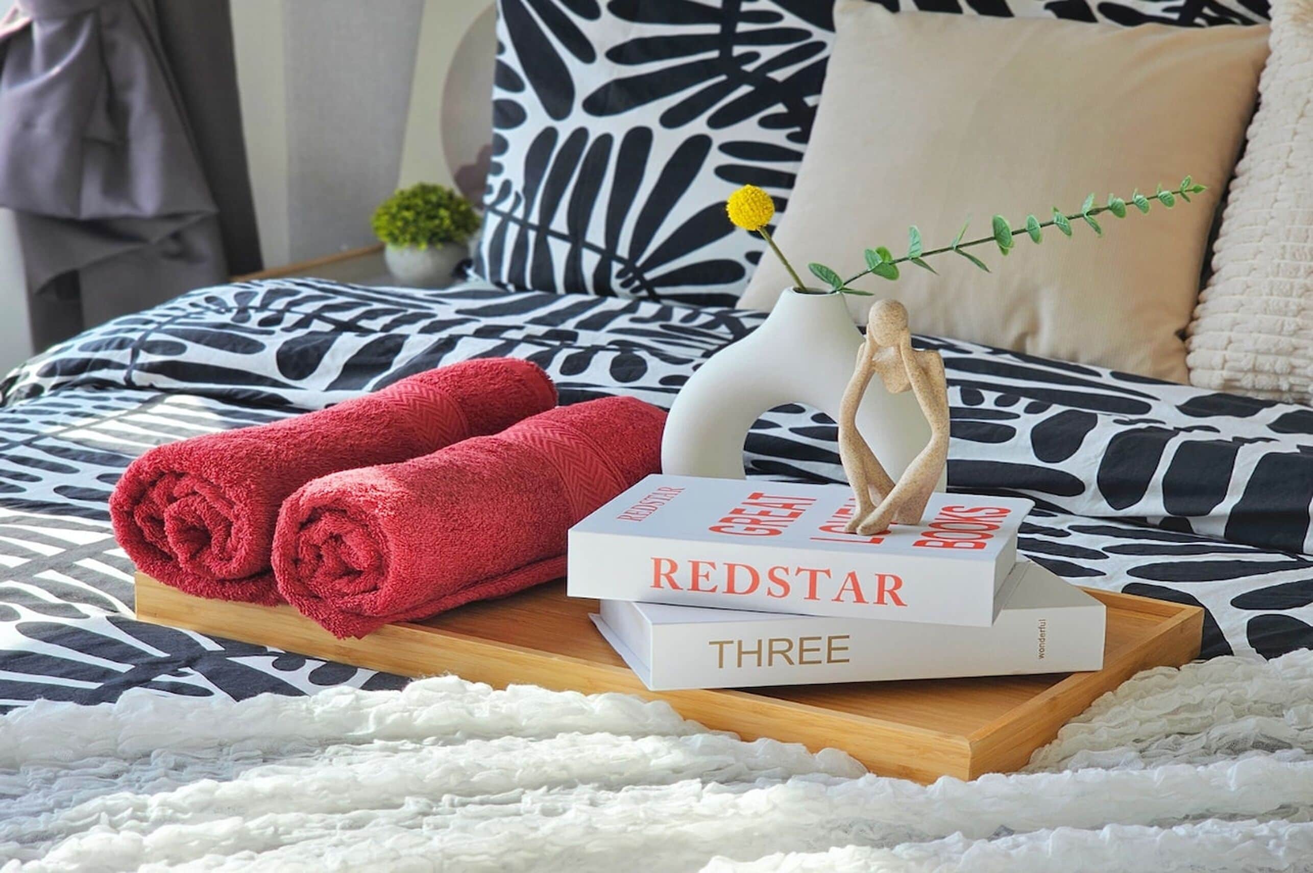A neatly made bed with a patterned bedspread, a small potted plant, and a tray with rolled towels and books.