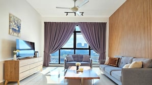 Una sala de estar moderna con un televisor de pantalla plana, un mueble de madera, un sofá gris, una mesa de centro y un ventanal con cortinas moradas.